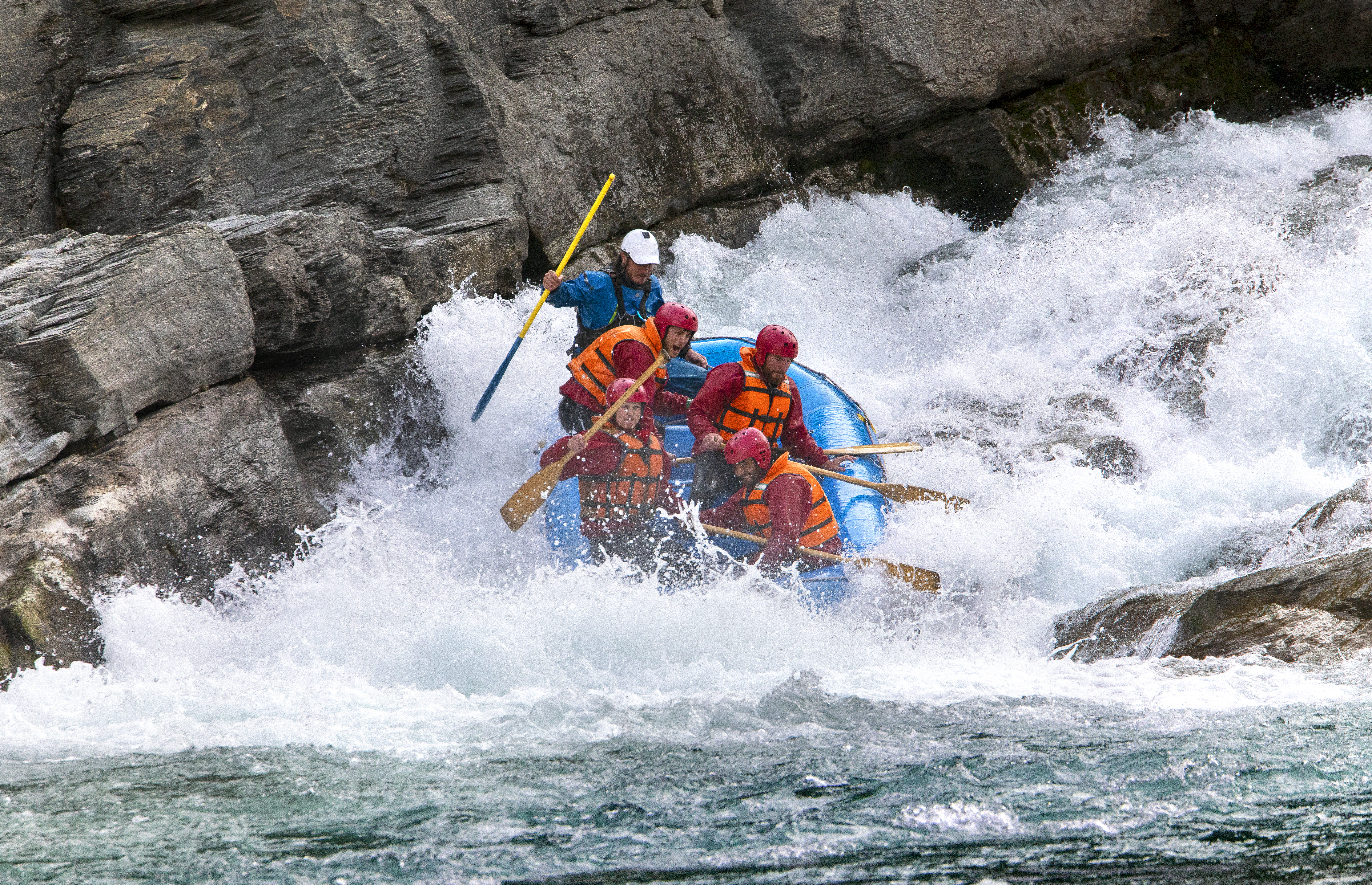 A group of people whitewater rafting through rapids, wearing helmets and life jackets, surrounded by rocky cliffs.